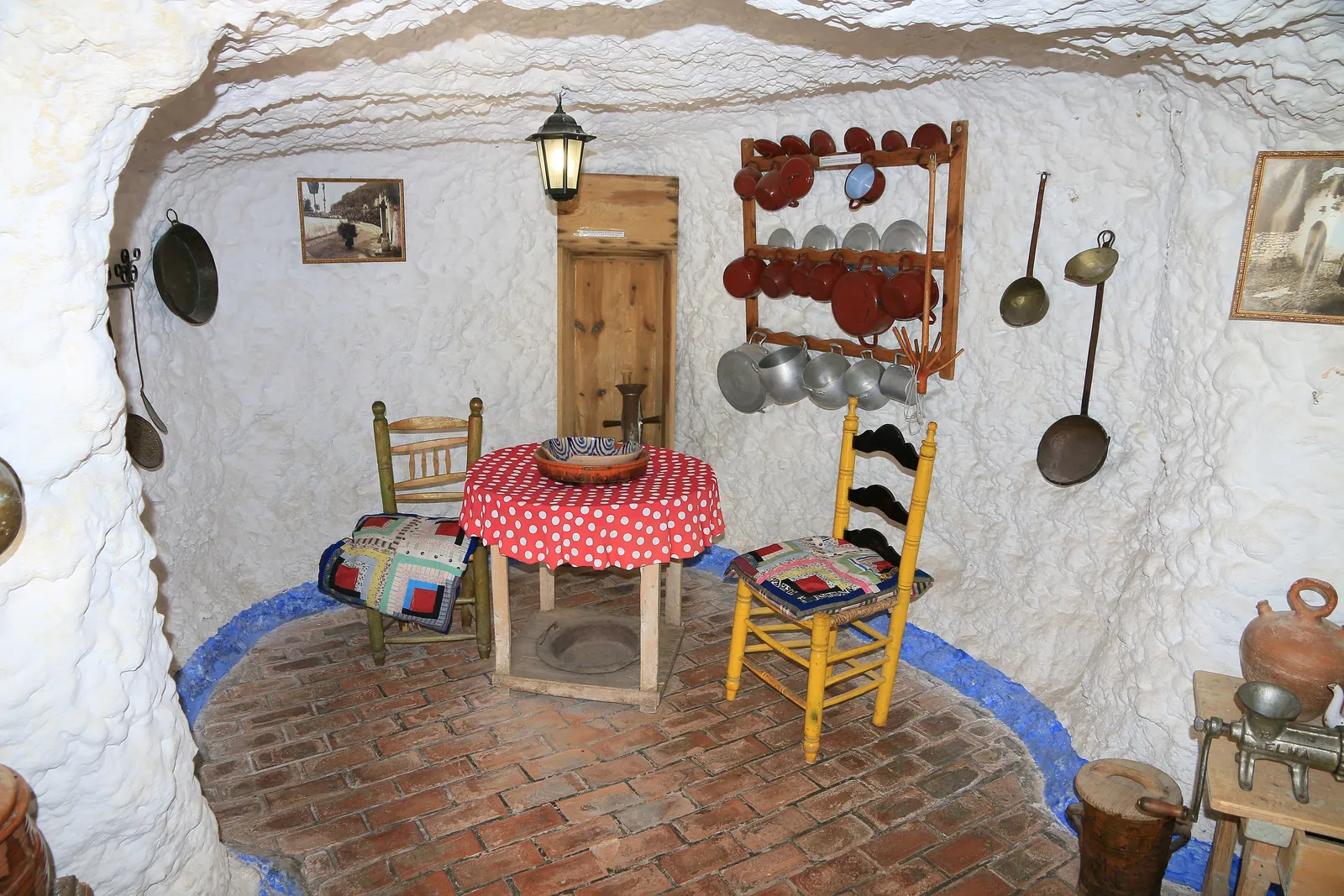 A whitewashed cave room with a red-spotted table, a yellow chair and copper pans on the wall, rebuilt at the Sacromonte Caves Museum; a general photograph of a cave home, not a show cave